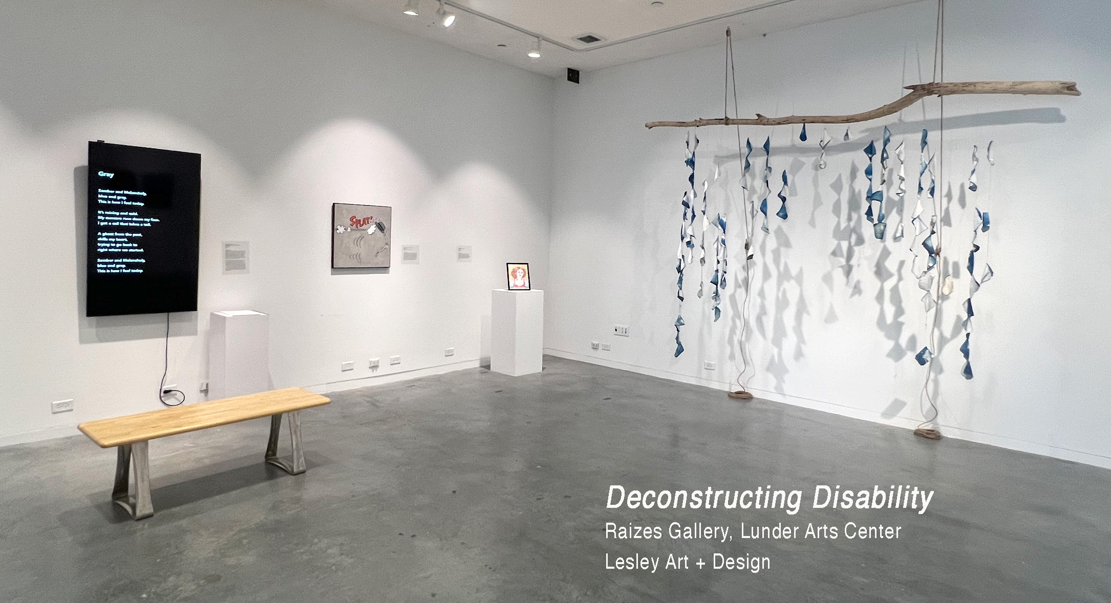 Deconstructing Disability exhibition photo. The photo is of a gallery with white walls and a gray floor. On the left wall is a monitor with a black background and a poem with white text. To the right of that is a square painting with cartoon imagery. In the center of the image and the corner of the wall is a framed painted self-portrait on a white pedestal. On the right wall is a sculptural work that is a large log hanging from the ceiling with strings of cyanotype photos that hang from the bottom of the log to the floor. These strings of images cover the width of the log.