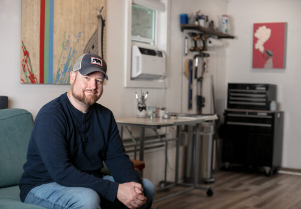 Photo of Bill Porter wearing a blue and white baseball cap, a Henley shirt, and jeans, sitting on a couch in the studio with his hands crossed on his lap. His tools and painting supplies are in the background, and his paintings are on the walls.
